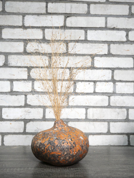 A rustic, flat, bulb-shaped gourd vase with a mottled orange and dark brown textyre, filled with dried ornamental grass, set against a white painted brick wall background.