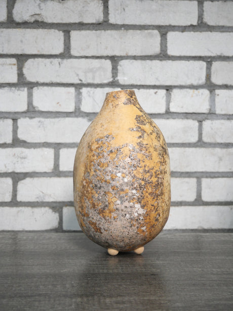 A handmade gourd vase with a rounded, bulbous shape and a narrow neck, featuring a textured patina in earthy brown, beige, and gray tones. It is displayed on a dark wooden surface against a white brick wall background.