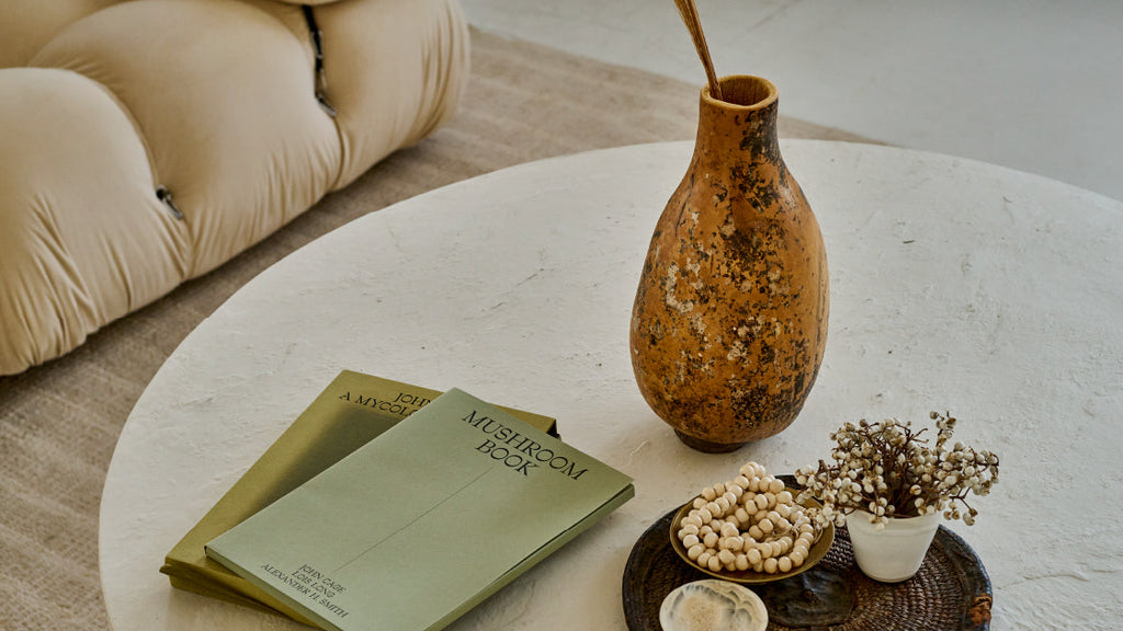 A styled interior scene showing a round white table with a tall speckled brown vase, a stack of green books, a small white pot with dried flowers, and a dark tray on a beige rug.