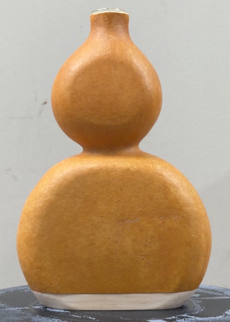 A natural dried gourd vase with a distinctive flattened double-bulb shape and a small neck, finished with a smooth matte finish in a warm golden-brown color, displayed on a dark surface against a light gray background.