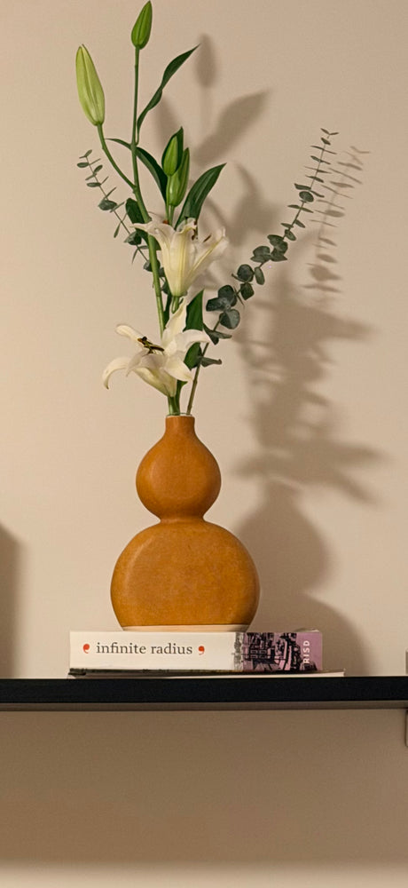 An orange-brown double-bulb gourd vase holding white lilies and eucalyptus, displayed on a shelf against a beige wall.